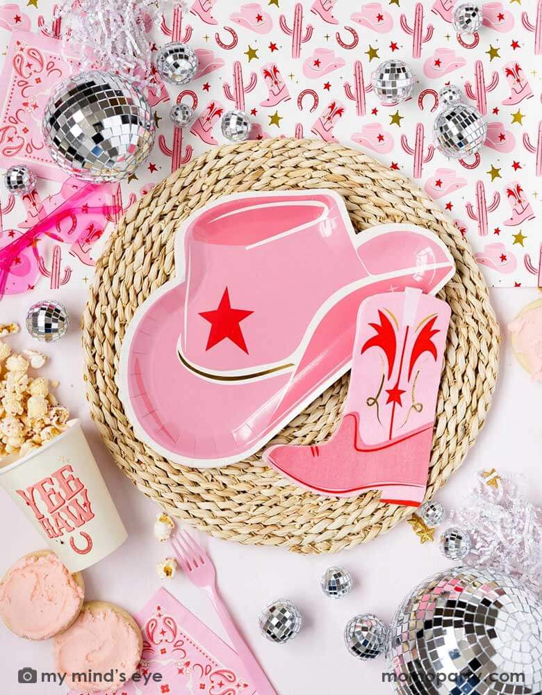 A cowgirl rodeo Western themed party table features pink cowgirl themed party supplies from Momo Party including pink cowgirl boot shaped napkins, cowgirl hat shaped plates, pink bandana small napkins, yeehaw party cups and cowgirl pattern paper table runner, along with festive disco ball decorations, heart shaped sunglasses and treats, this makes a perfect inspo for kid's cowgirl themed birthday party or a bachelorette party celebration!