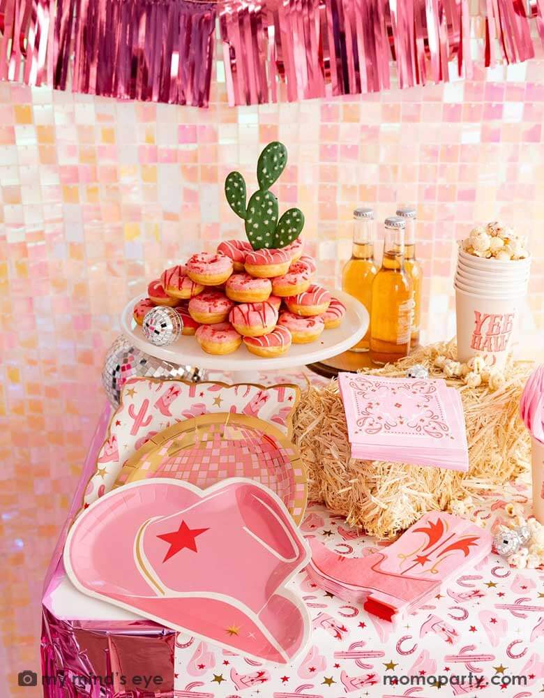 A cowgirl rodeo Western themed party table features pink cowgirl themed party supplies from Momo Party including pink cowgirl boot shaped napkins, cowgirl hat shaped plates, pink disco ball shaped plates, pink bandana small napkins, yeehaw party cups and cowgirl pattern paper plates and table runner by My Mind's Eye. In the back there's an iridescent backdrop adorned with pink foil fringe banner, along with pink donut tower cake topped with a cactus, making this a great inspo for a disco cowgirl party.