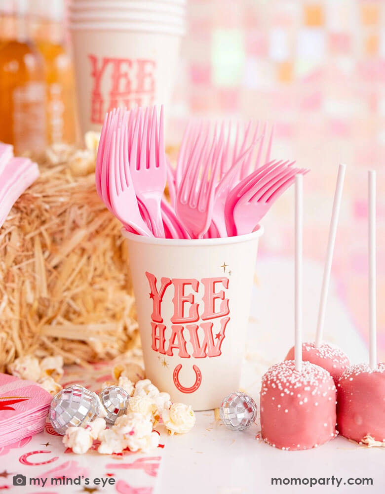 A cowgirl rodeo Western themed party table features pink cowgirl themed party supplies from Momo Party including 12oz pink YEEHAW party cups with pink utensils, cowgirl boot shaped napkins on the cowgirl pattern paper table runner, along with hay stacks, some small disco ball decorations and pink cake pops and popcorn, making this a perfect inspo for kid's cowgirl themed birthday party or a disco cowgirl bachelorette party celebration!