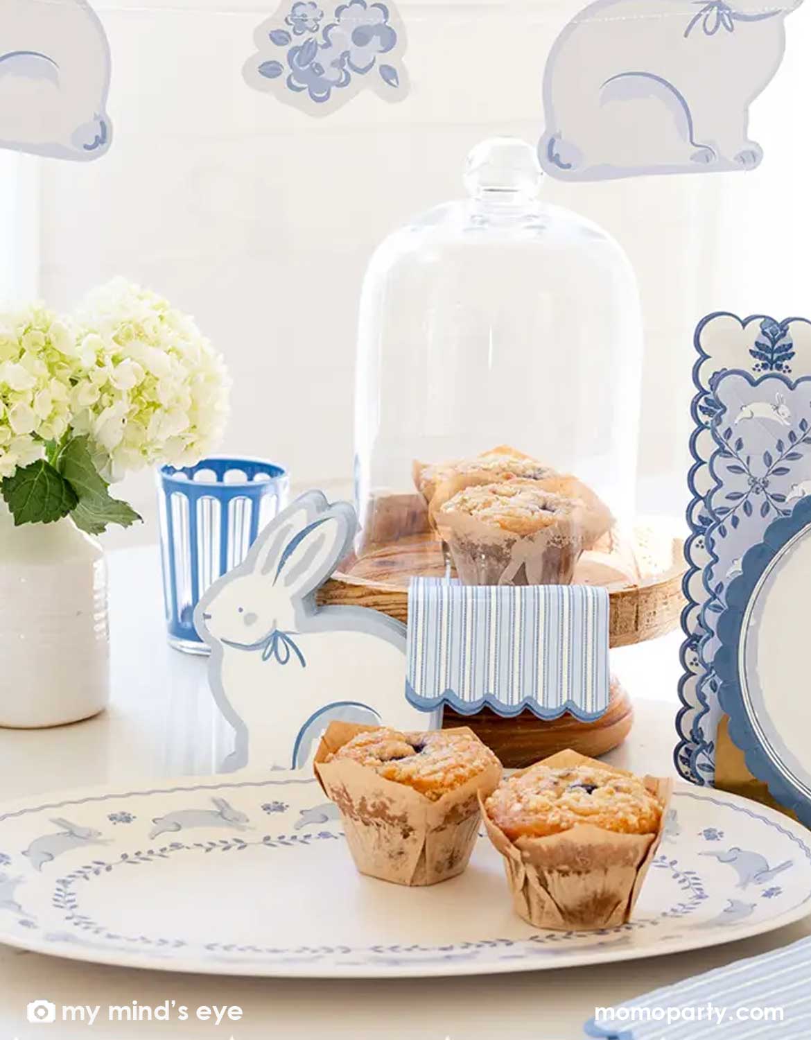 An elegant Easter brunch table setting featuring multiple blue bunny themed tablewares from Momo Party including the blue oval bunnies bamboo serving tray with muffins on it, blue bunny shaped napkins, blue striped guest napkins, bunnies pattern square plates and blue floral scallop round plate. With spring flowers around and bunny shaped banner hung above, this makes a great inspo for a beautiful Easter table setting idea.