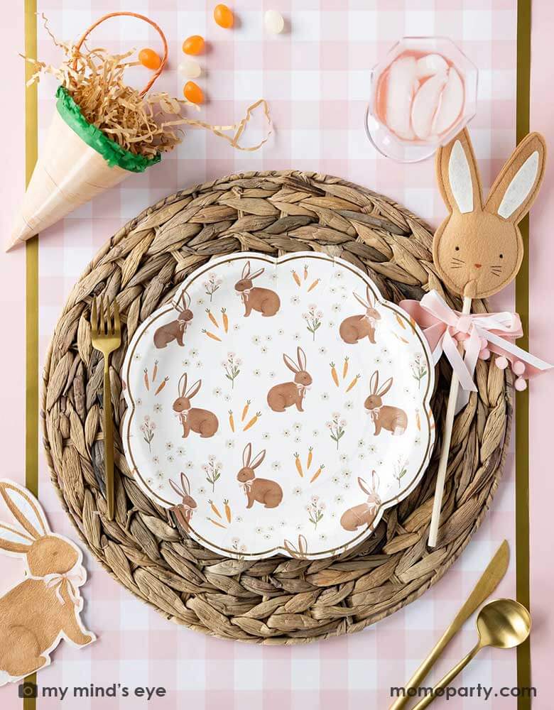 A beautiful Easter tablescape features Momo Party's scatter rabbit scallop paper plate by My Mind's Eye in the middle, laid on top of a straw woven placemat which is on the pink gingham checks paper table runner, around the plate are carrot shaped treat boxes, brown rabbit shaped napkin and Easter rabbit wand, makes this an adorable kid's friendly Easter tableset this spring.