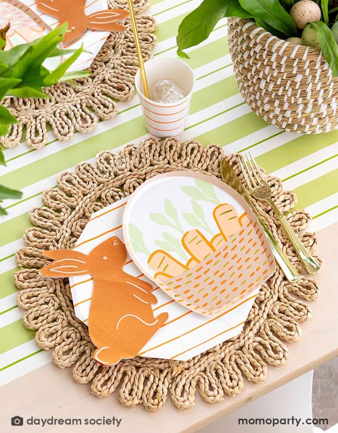 An Easter bunny themed table setting with Momo Party's bunny themed tablewares including carrot basket plates, bunny shaped napkins and matching cups and utensils on a straw-weaved placemats over a green and white striped table runner.