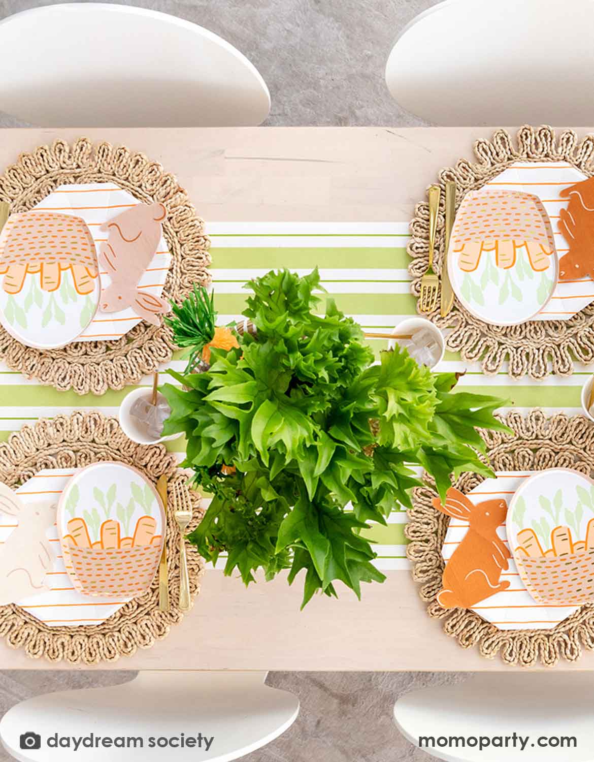 An overhead shot of an Easter bunny themed table setting with Momo Party's bunny themed tablewares including carrot basket plates, bunny shaped napkins and matching cups and utensils on a straw-weaved placemats over a green and white striped table runner. In the center there's a green and carrot floral arrangement as the centerpiece.