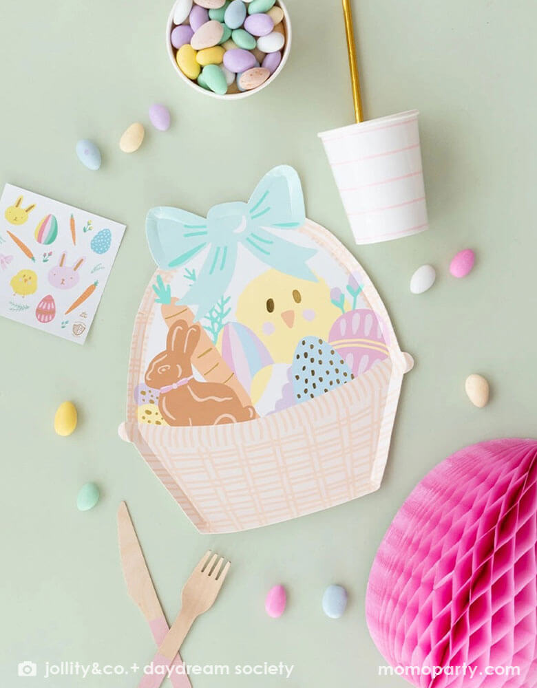 A pale mint table features Momo Party's 9.5 x 10.75 inches Easter basket shaped large plates by Daydream Society. Around it are some easter egg shaped candies, a light pink striped party cup, an Easter Fun sticker, pink wooden cutlery and sheet and a pink Easter egg tissue honeycomb.