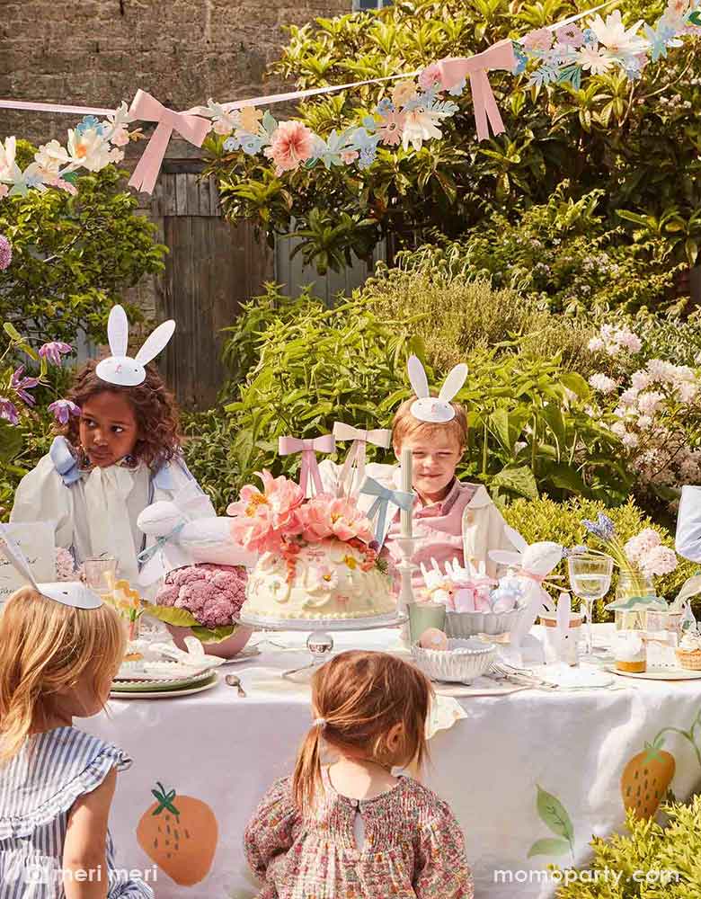 An adorable Easter party in the garden for kids featuring a long party table filled with Easter themed party supplies including bunny party cups, napkins, plates and party crackers. In the middle of the table, there's a large cake adorned with floral toppers and Momo Party's 3D bow cake toppers in pastel colors. Above the table hung the flowers and bows party garland, creating a wow factor to this gorgeous Easter celebration set up.