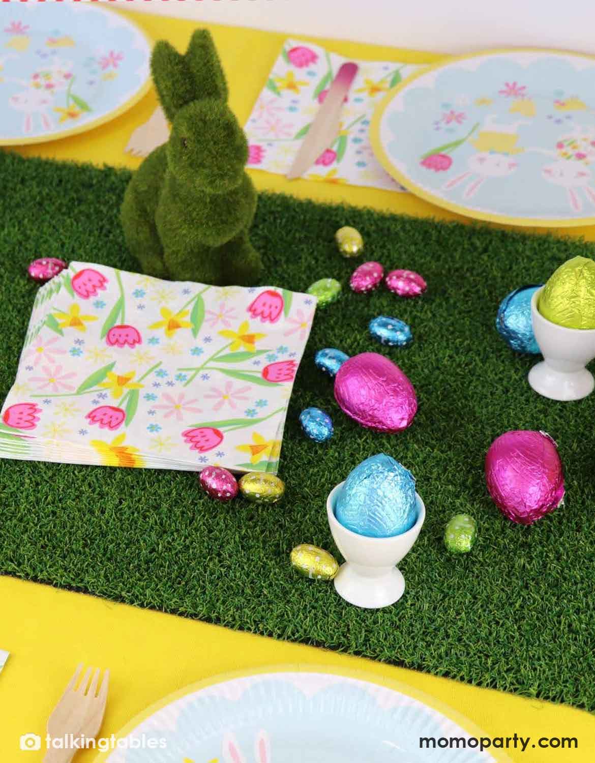 A styled Easter table setting with a green artificial grass table runner, pastel floral plates, a green bunny decoration, foil-wrapped chocolate eggs in white egg cups, and a floral napkin on a yellow tablecloth.