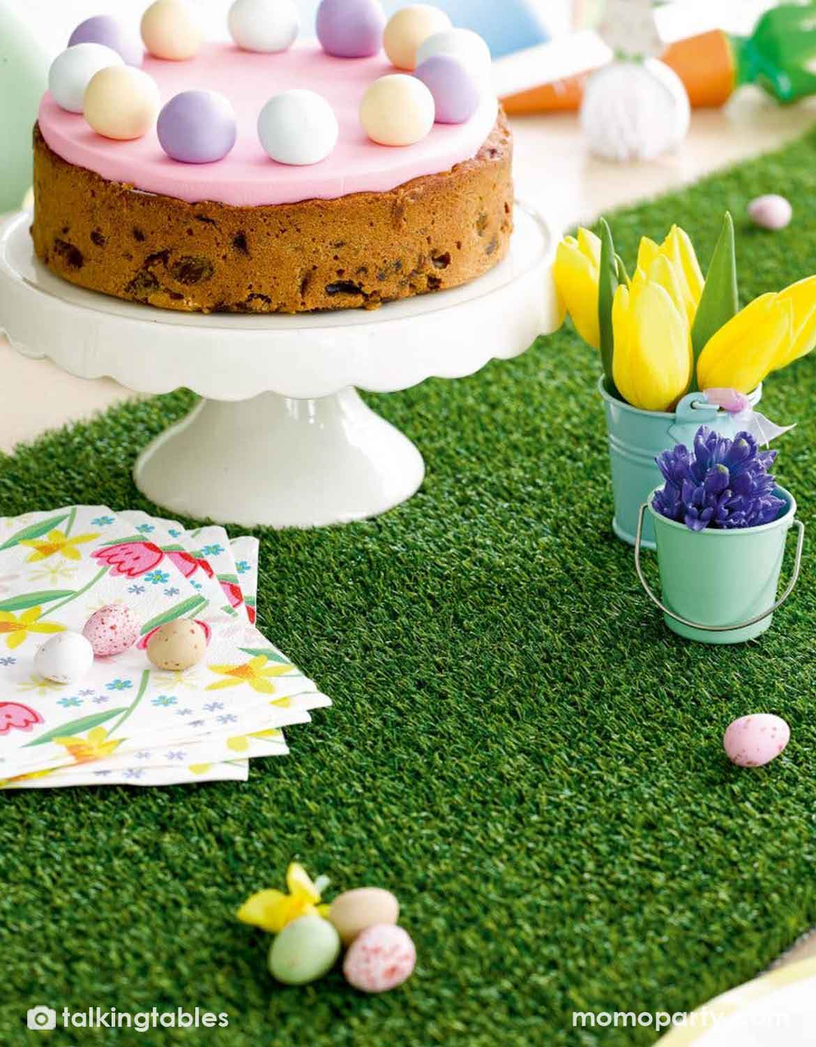 An Easter-themed scene with a round cake topped with pastel eggs on a white pedestal stand, set on artificial grass table runner with tulips, pastel eggs, napkins, and small decorative buckets.
