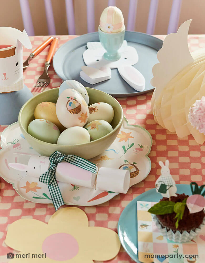 An Easter party table with Easter eggs adorned with Meri Meri's Easter themed tattoo stickers, on the table are Momo Party's Easter themed plates and napkins in soft pastel colors including bunny shaped napkins and party cups and bunny party crackers which are coordinated with the charming daisy shaped plates, makes this an adorable tablescape idea for a spring gathering.