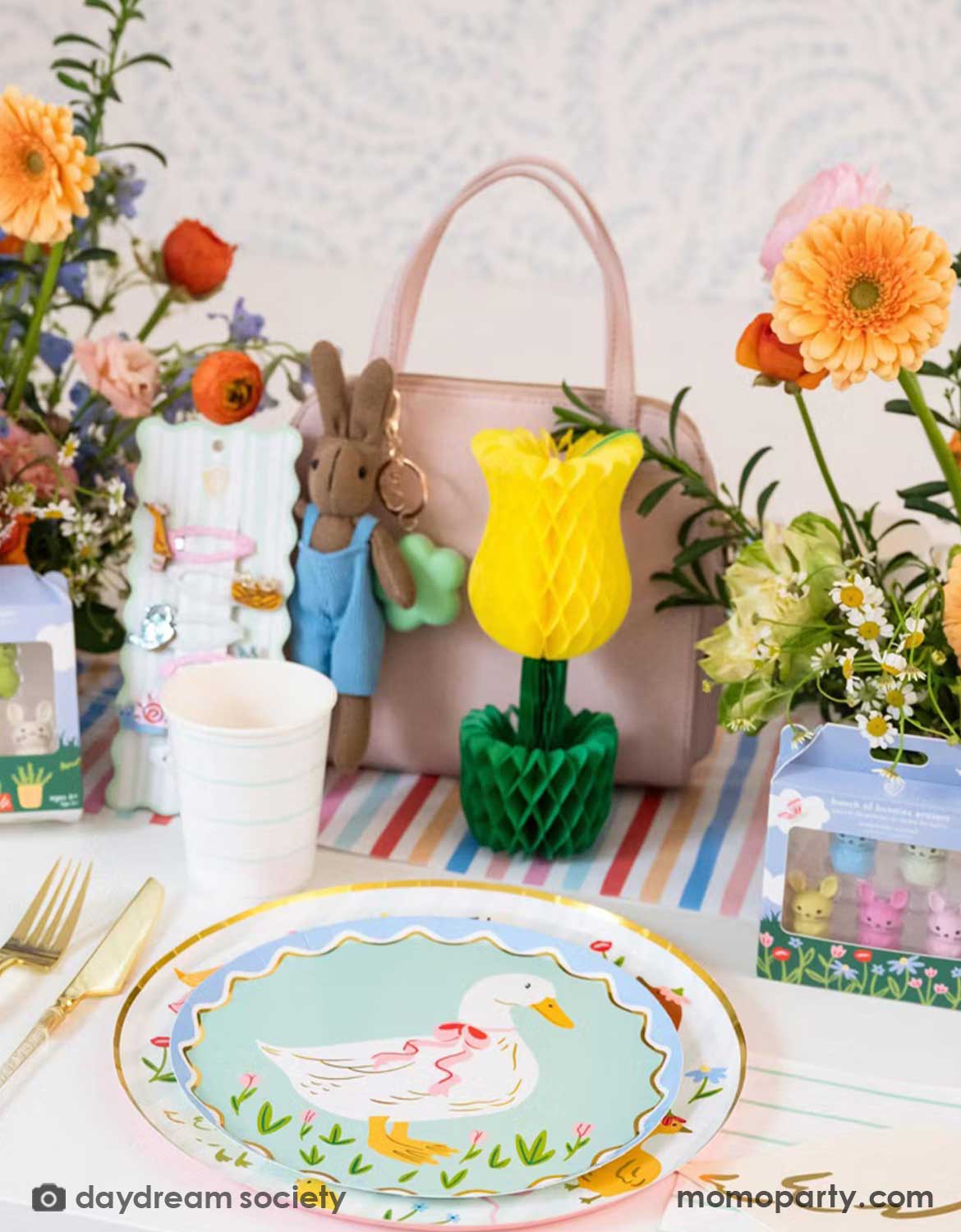 A children's Easter table setting with colorful flowers, tulip honeycomb decoration, goose themed party plates, napkins, party cups, cutlery and some kid's Easter small gifts with bunny designs on a colorful striped runner. A perfect idea for an Easter celebration, a baby shower, kid's Silly Goose themed birthday party or a spring gathering.