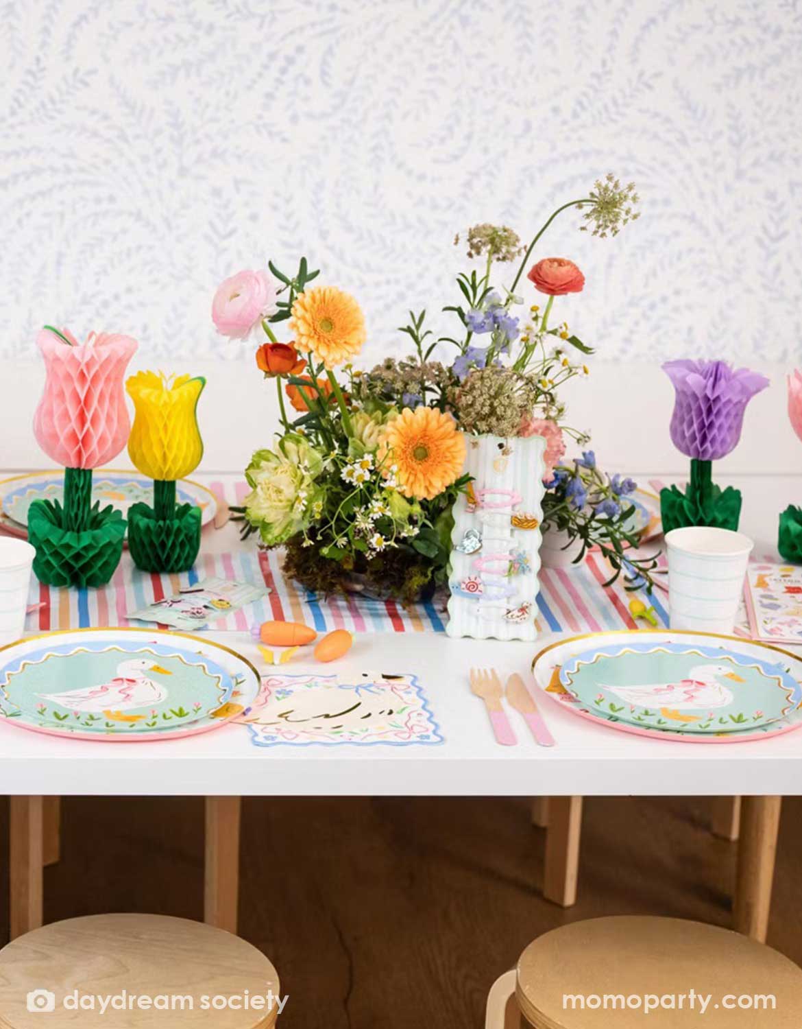 A children's Easter, spring themed table setting with colorful flowers, tulip honeycomb decorations, goose themed party plates, napkins, party cups and cutlery on a colorful striped runner. A perfect idea for an Easter celebration, a baby shower, kid's Silly Goose themed birthday party or a spring gathering.