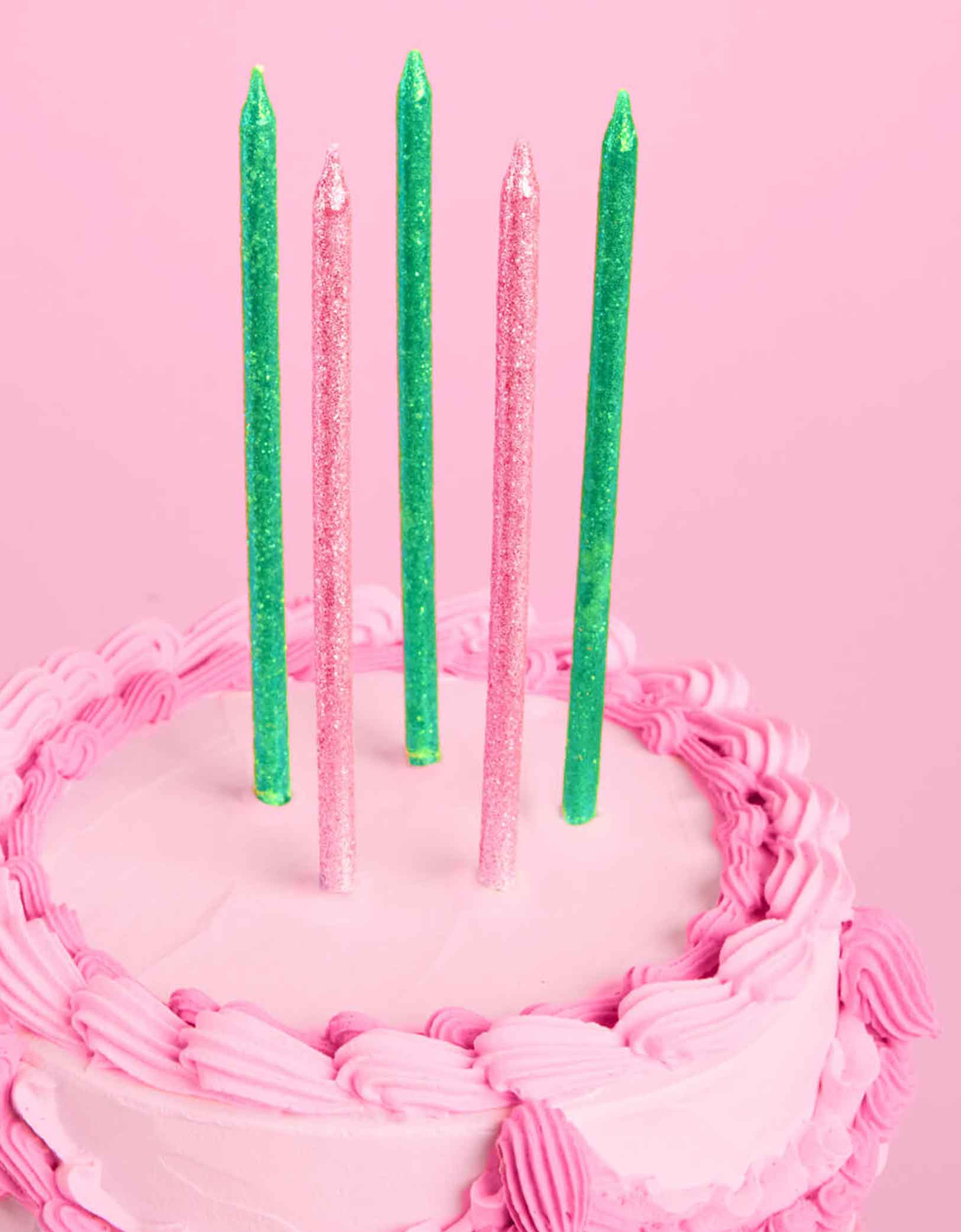 A pink buttercream cake with Momo Party's Wicked Party Emerald City Glitter Candles in pink and green by Xo, Fetti on a pink background.
