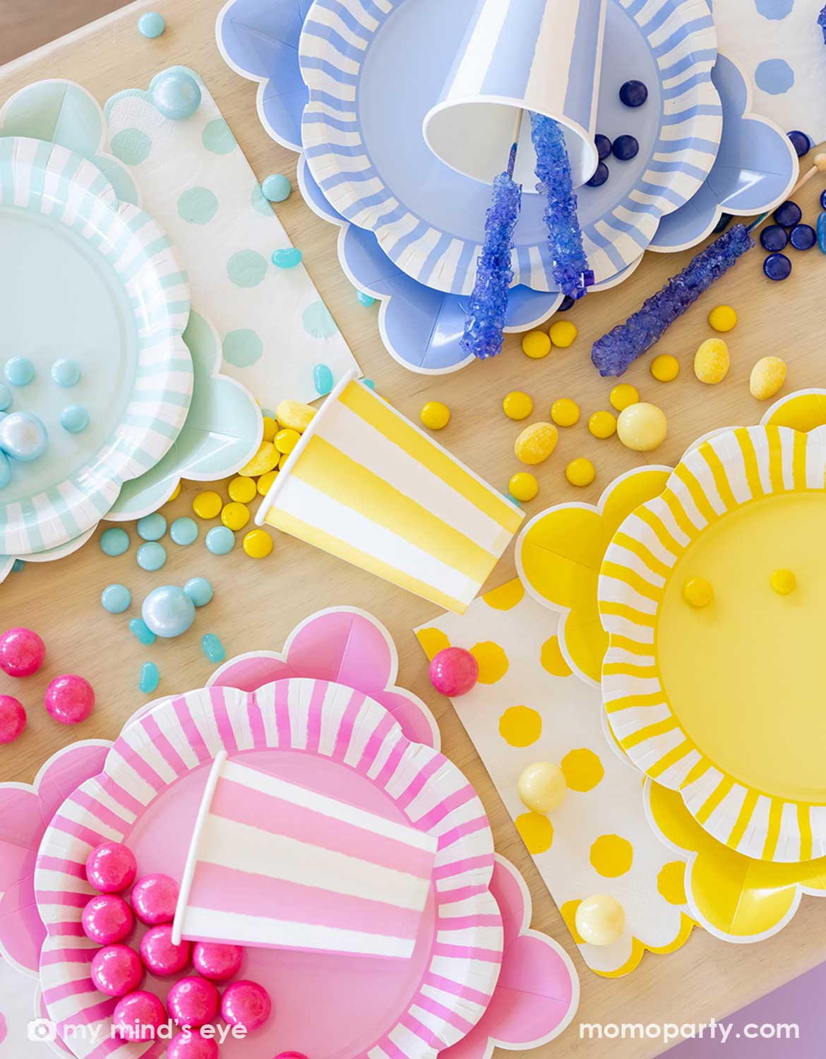 An overhead shot of Momo Party's colorful paper plates, cups and napkins with color-coordinated decorative elements and treats in yellow, pink, blue and mint colors on a wooden table.