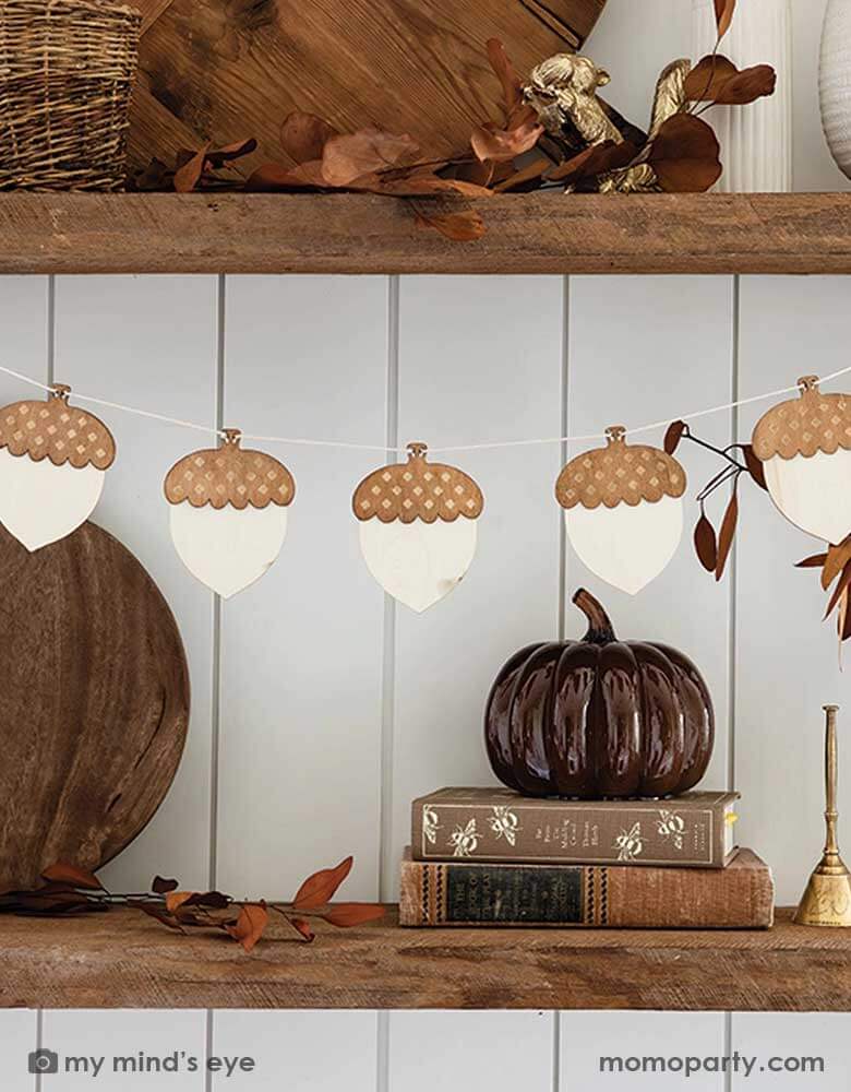 A fall inspired kitchen shelf decorated with Momo Party's 9ft wood acorn banner by My Mind's Eye. On the shelf, there are some fall colored table decorations including wooden cutting boards, books in brown covers, gold metal squirrel and broom decoration, some baskets and fall leaves spread around - a perfect inspo for fall home decors in this harvest season.