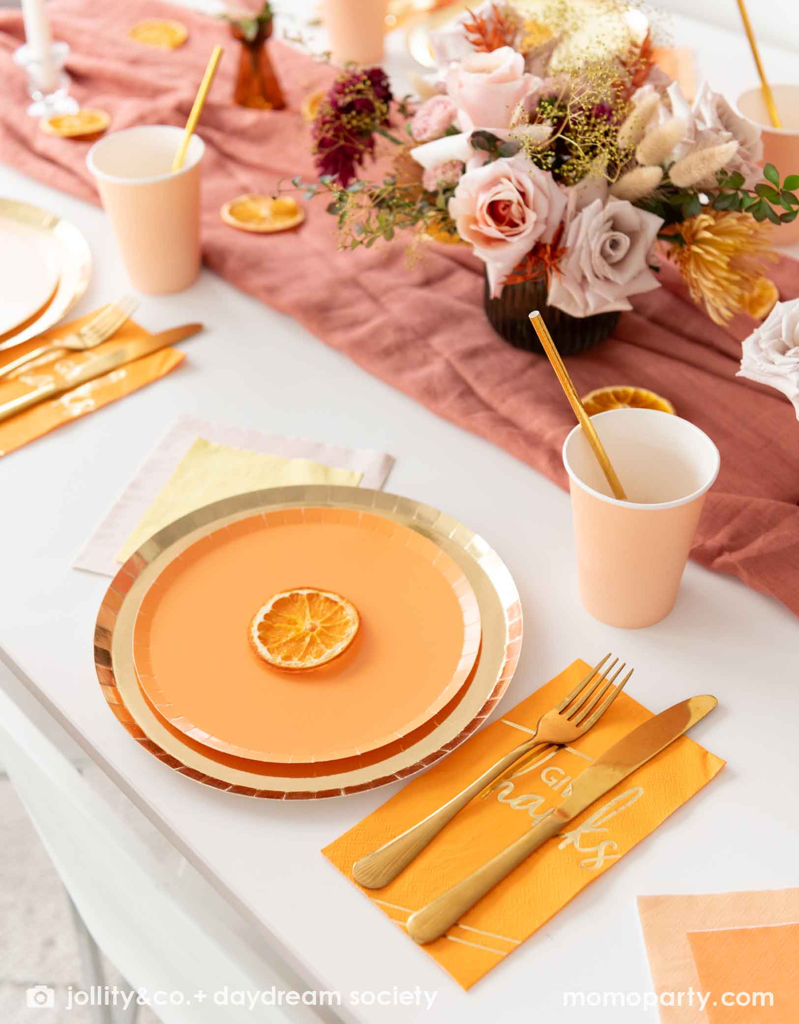 A fall inspired decorative table setting with Momo Party's apricot plates, and gold plates, peach cups with orange guest napkins with thankful text on them and gold cutlery. In the center there's a fall themed floral bouquet with orange slices spread out.
