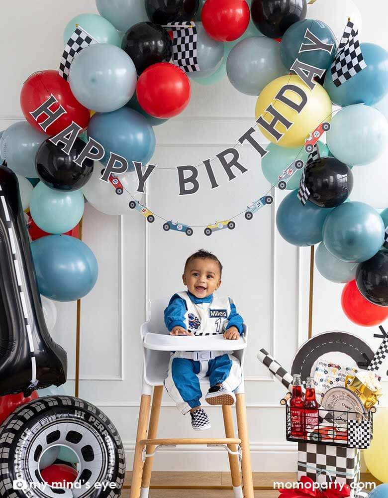 A one-year-old boy in his racer jumper sitting in his high chair in a "Fast One" "Number One" race car themed first birthday party set up decorated with a colorful balloon garland, race car happy birthday banner, a number 1 foil balloon decorated with road tape, a tire shaped foil balloon and a basket of race car themed tableware including race car plates, napkins, party cups, checkered table runner and trophy treat boxes. Making this a perfect inspo for boy's first birthday party decoration.
