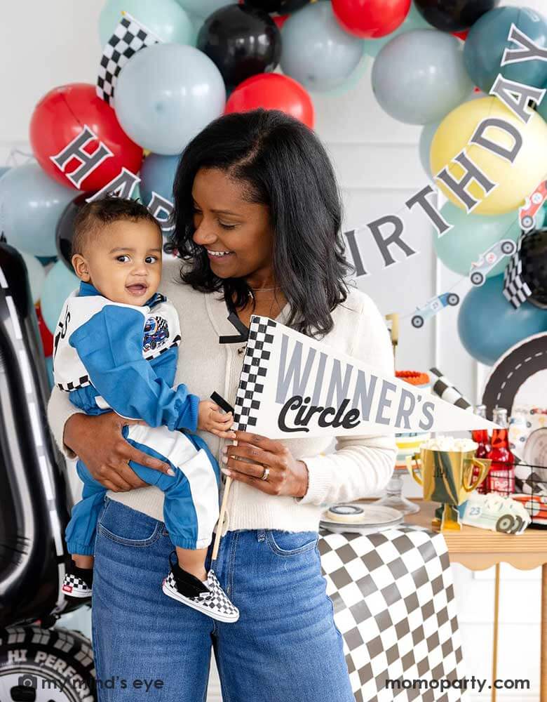 A mom holding his one year old boy in her arm standing in front of a race car themed party decoration set up which includes a colorful balloon garland in red, blue, gray black and yellow adorned with a happy birthday paper banner, on the table there are race car themed partyware including plates, napkins, checkered table runner and race car flags. The mom is holding a felt party pennant written "Winner's Circle" from Momo Party.
