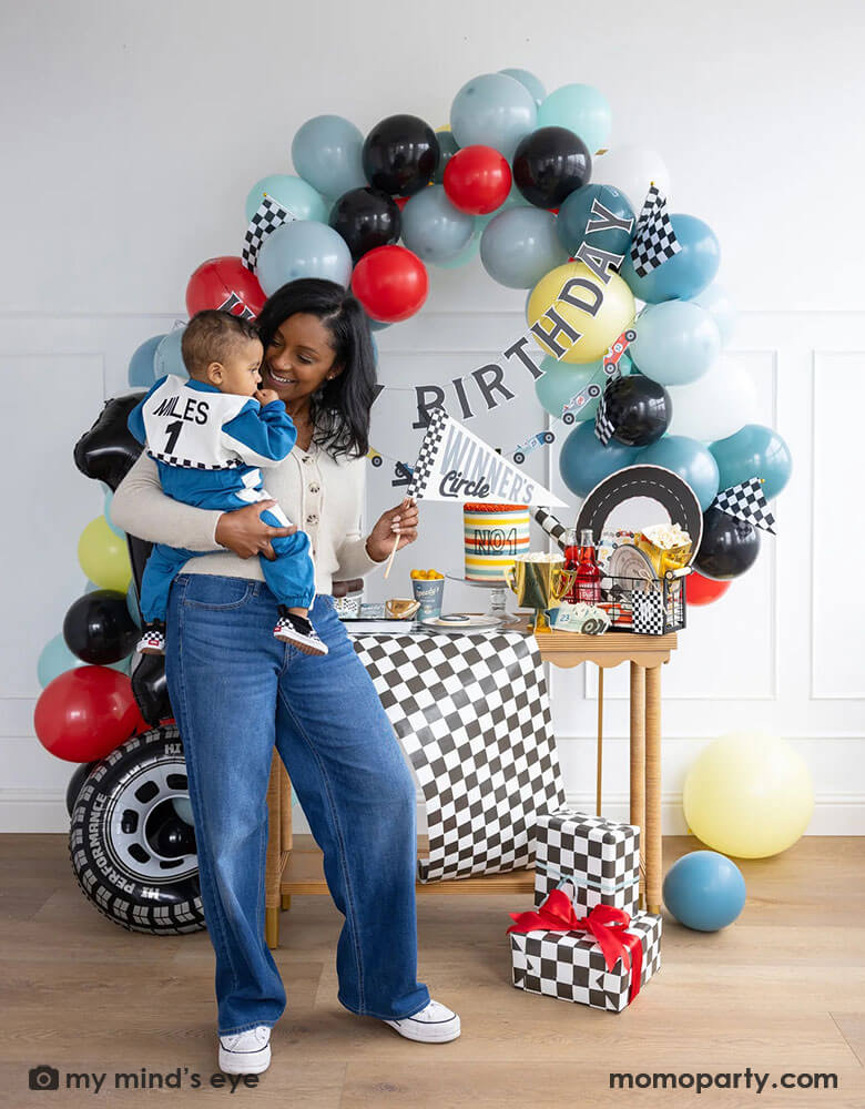 A mom holding his one year old boy in her arm standing in front of a race car themed party decoration set up which includes a colorful balloon garland in red, blue, gray black and yellow adorned with a happy birthday paper banner, on the table there are race car themed partyware including plates, napkins, checkered table runner and race car flags. The mom is holding a felt party pennant written "Winner's Circle" from Momo Party. On the floor there are some birthday gifts wrapped with checkered paper.
