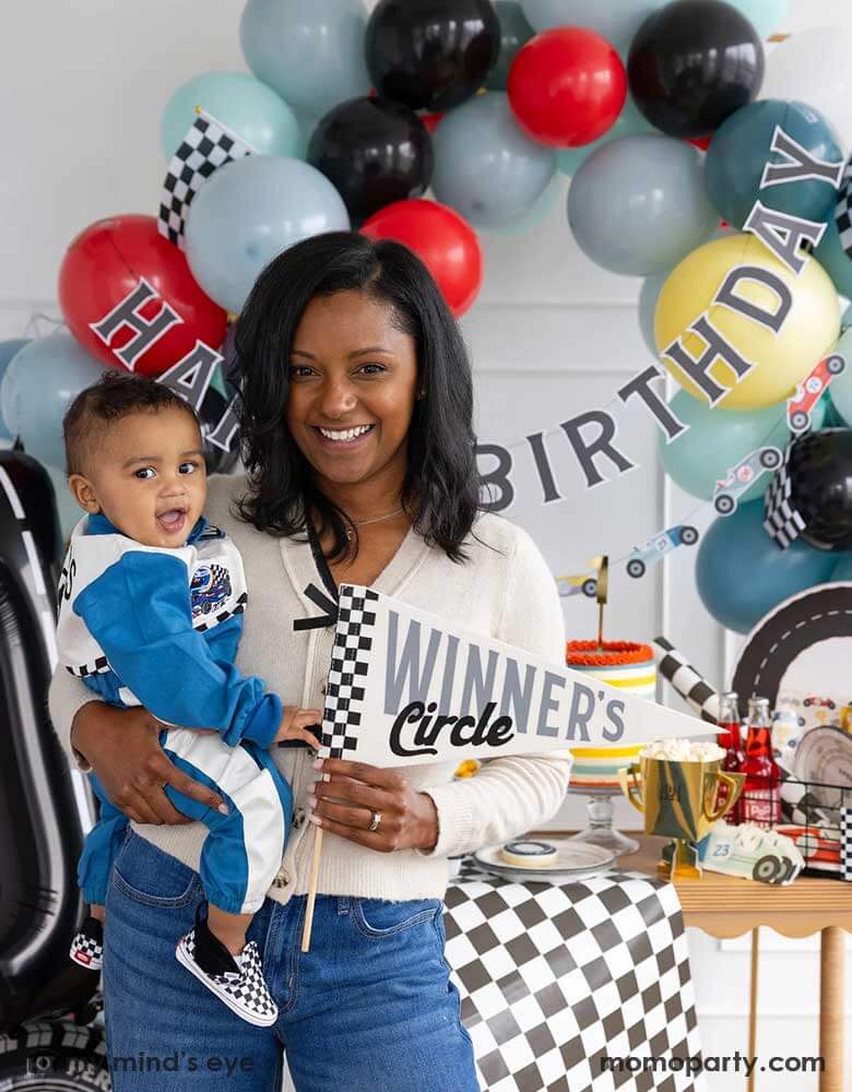 A mom holding his one year old boy in her arm standing in front of a race car themed party decoration set up which includes a colorful balloon garland in red, blue, gray black and yellow adorned with a happy birthday paper banner, on the table there are race car themed partyware including plates, napkins, checkered table runner and race car flags. The mom is holding a felt party pennant written "Winner's Circle" from Momo Party.