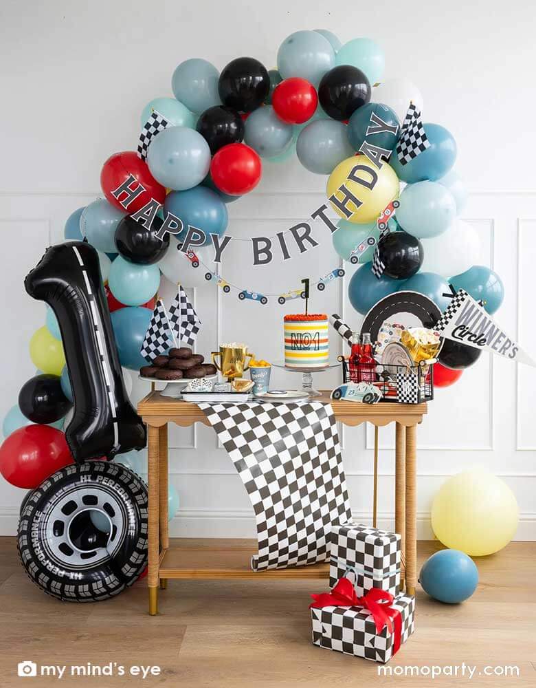 A "Fast One" boy's race car themed first birthday party decoration featuring festive race car themed party supplies from Momo Party including Miles per Hour party plates, cups, napkins, placemats, trophy treat cups, and a checkered table runner. On the party table there are a cake and donuts topped with checkered flags. Above the table is an organic balloon garland adorned with race car happy birthday banners. Along with a tire shaped foil balloon a number one letter balloon with decorated with road tape.