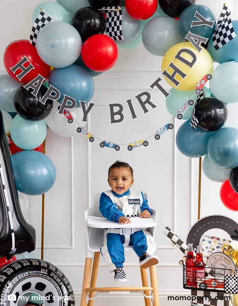 A one-year-old boy sitting in his high chair in a "Fast One" "Number One" race car themed first birthday party set up decorated with a colorful balloon garland, race car happy birthday banner, a number 1 foil balloon decorated with road tape, a tire shaped foil balloon and a basket of race car themed tableware including speedometer shaped plates, race car shaped napkins, checkered table runner and trophy shaped treat boxes. Making this a perfect inspo for boy's first birthday party decoration.