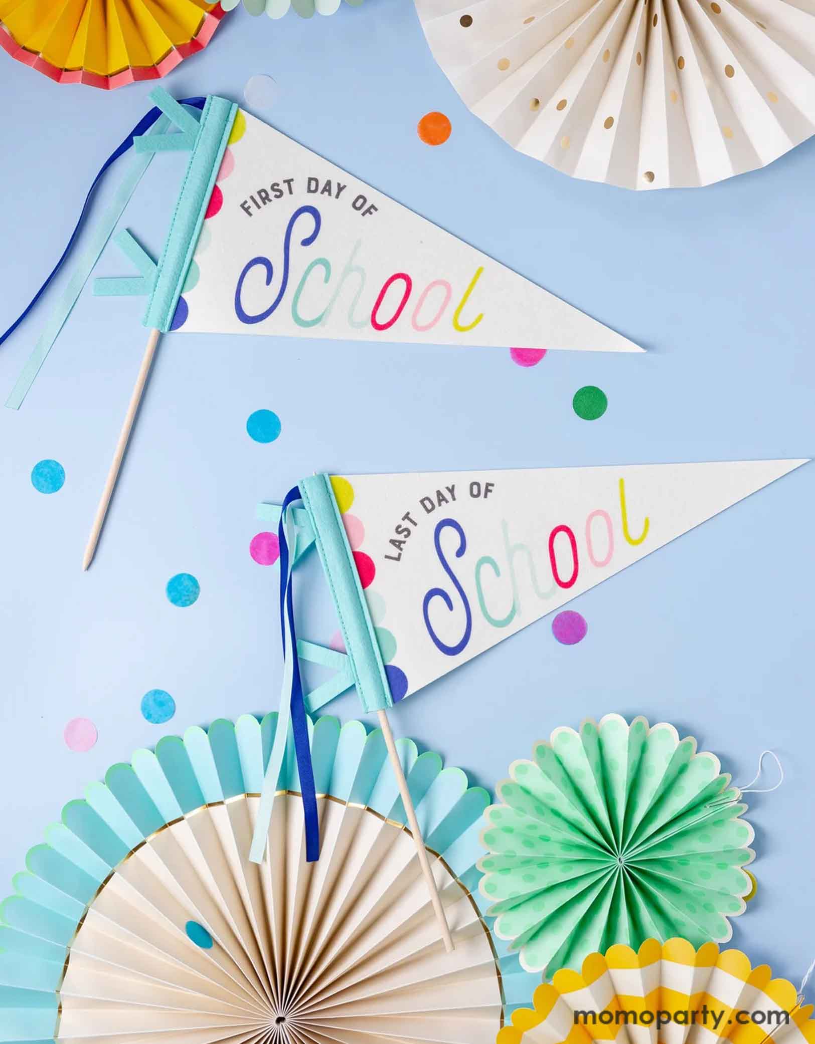 An overhead shot of Momo Party's 14" First Day of School/Last Day of School Pennant Banner Set by My Mind's Eye on a blue background. Around the pennants are colorful paper fans and round confetti.  This playful set features pennants for the first and last day of school, adding a unique touch to your classroom or home decor. Get ready to celebrate and capture those special moments with these festive and fun banners.