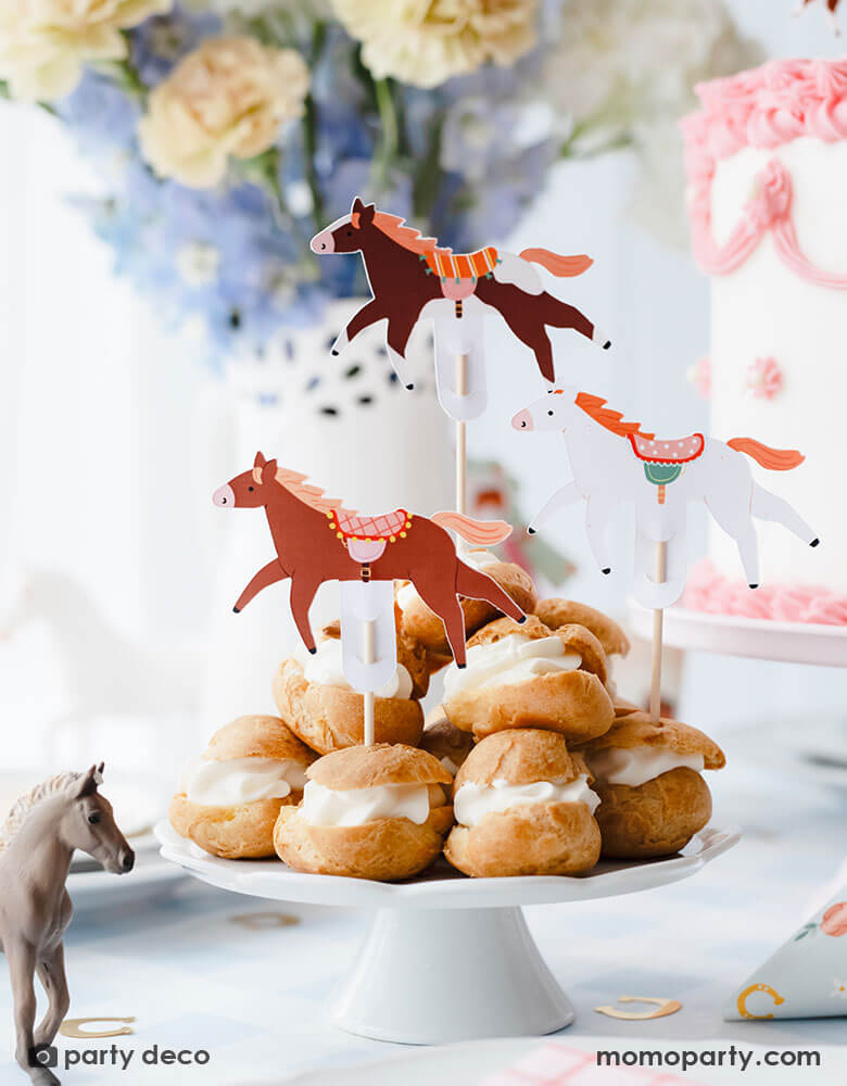 A beautiful horse themed party table featuring some delicious cream puffs stacked topped with Momo Party's floral pony cupcake toppers in 3 different colors and designs. On the table there are some gold horseshoe shaped confetti, a pink and white buttercream cake and a beautiful bouquet in lilac and cream colors, with the horse figurine as the centerpiece, this gives a charming and darling idea for kid's pony or horse themed birthday party.