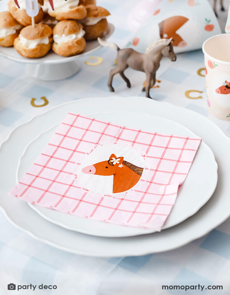 A pony horse themed birthday party table place setting featuring Momo Party's floral pony themed party supplies including the blue gingham tablecloth, large floral pony pink gingham napkins, party cups, and gold horseshoe shaped confetti around. On the table you can see some cream puff stacked in the back as dessert and a small horse figurine as the centerpiece - a great inspiration for kid's pony or horse themed party decoration.
