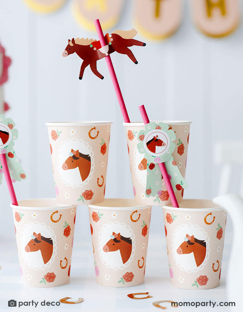 A stack of Momo Party's 6.7 oz floral pony party cups featuring adorable pony, floral and horseshoe design in blush color. There are also the floral pony paper straws in pink with paper pennants. On the table you can see some gold horseshoe shaped confetti, making this a perfect inspiration for kid's pony or horse themed birthday party set up.