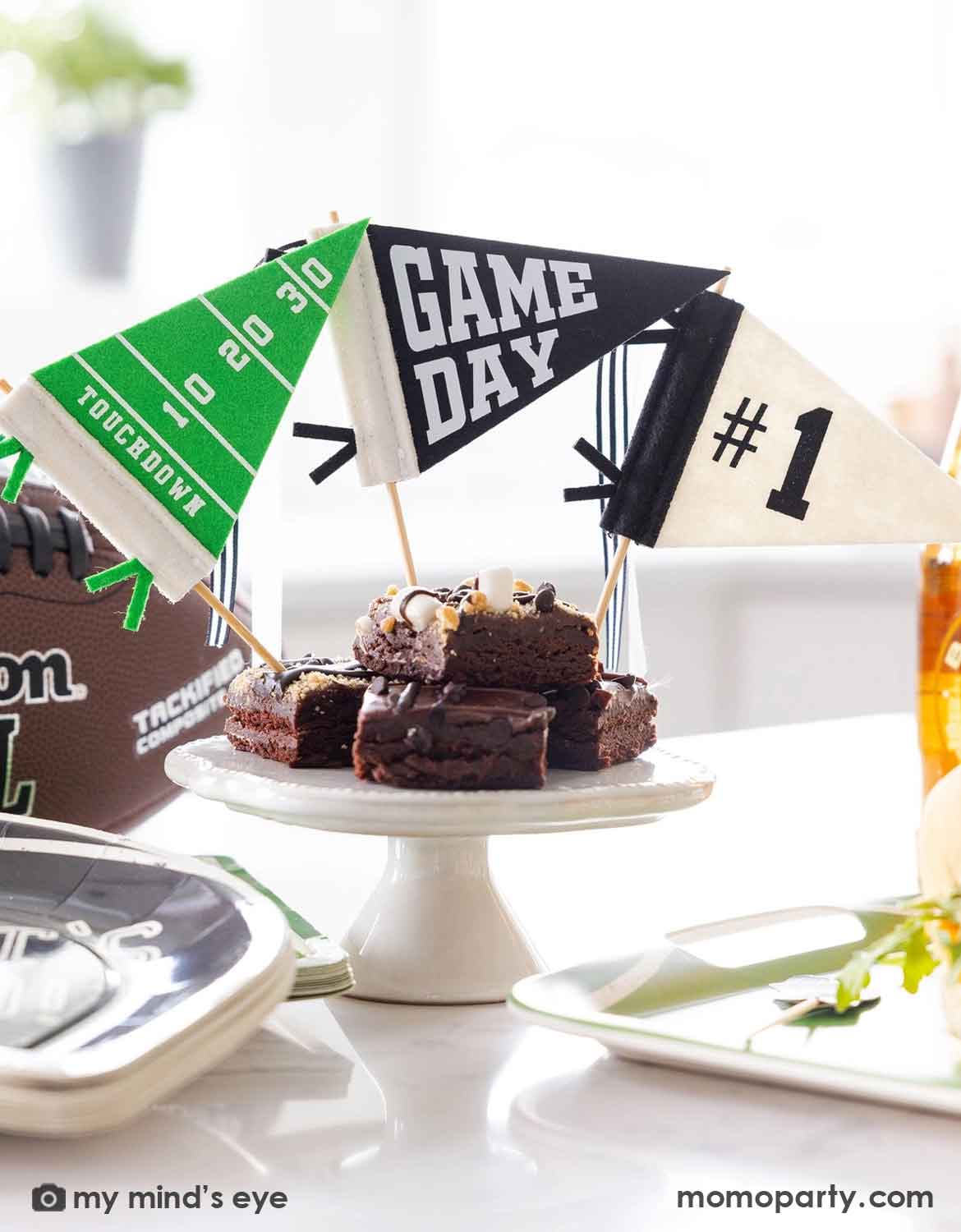 Football-themed party decorations and treats on a white table, featuring a brown football-shaped cupcake topper, mini game day flags reading 'GAME DAY', '#1', and 'TOUCHDOWN', and chocolate brownies on a white pedestal stand.