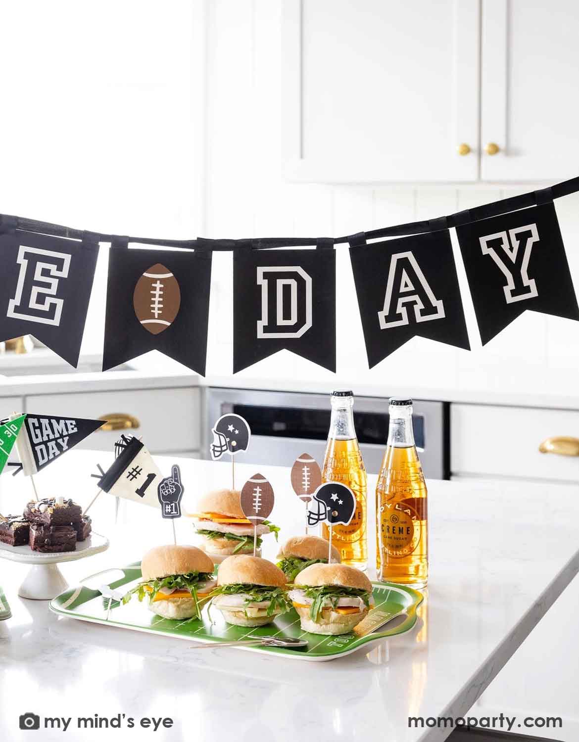 A football-themed party setup in a bright kitchen, featuring a black bunting banner that spells "Game Day" with football motifs, mini hamburgers on a green tray, two bottles of beer, and small 'GAME DAY' flags.