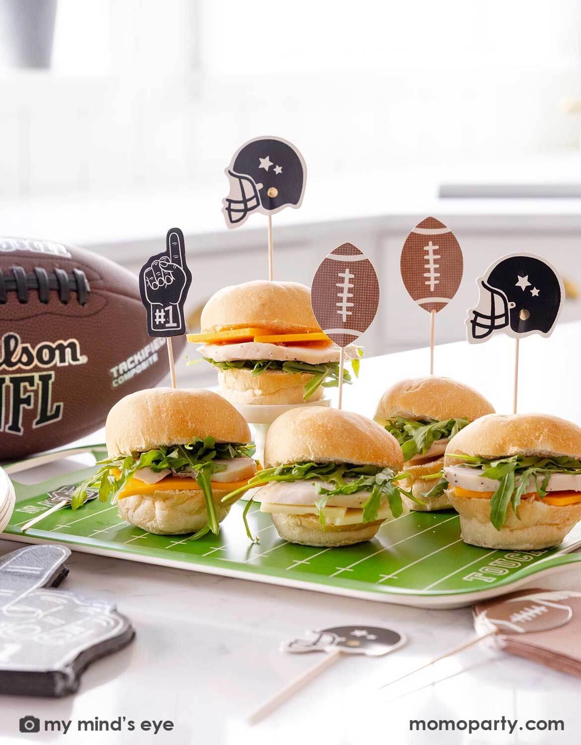 A football themed party with mini sandwiches on a football-themed tray  with football themed food picks from Momo Party and a football in the background.