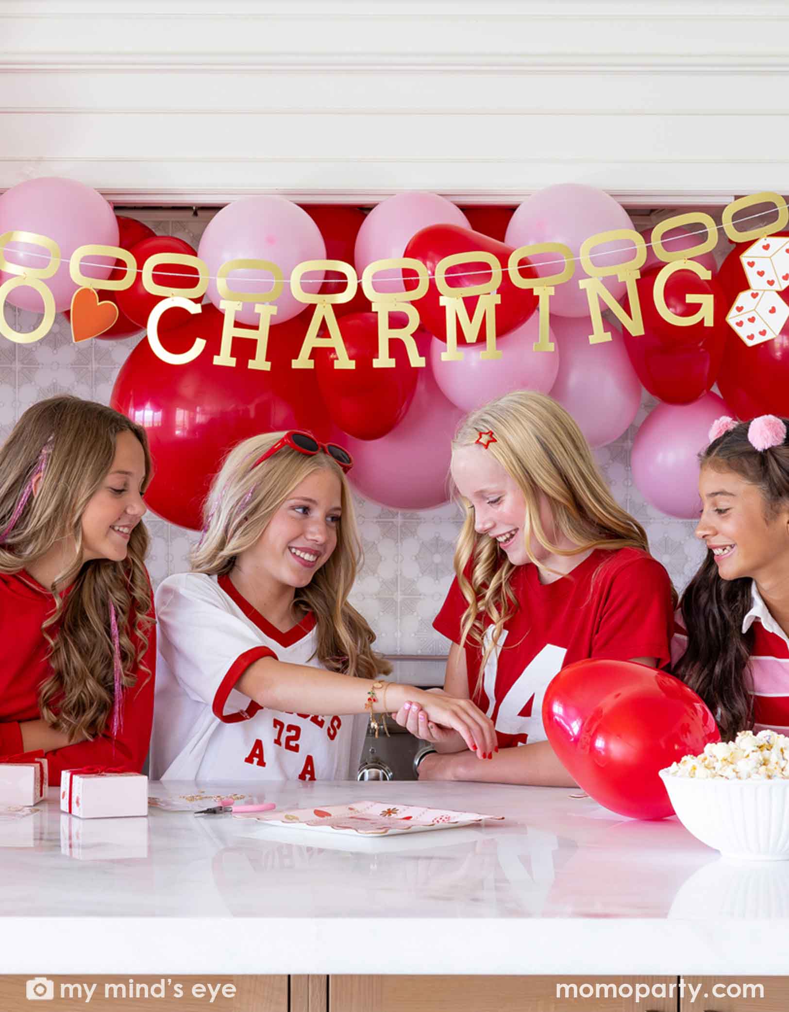 Four girls sitting at a Valentine's Day party table filled with heart themed party supplies from Momo Party including plates, napkins, and a cake. In the back there's a heart-themed balloon arch in red and pink and a gold foil party banner spells "So Charming". 