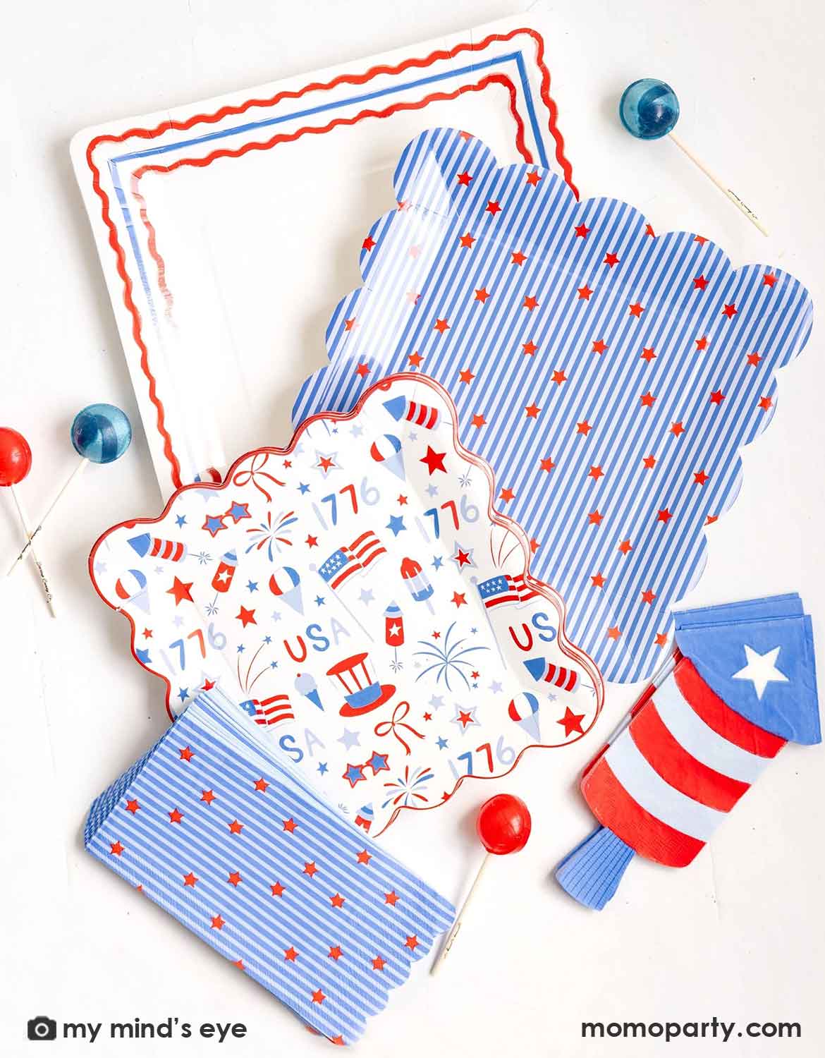 An overhead shot of USA themed tablewares from Momo Party including paper plates with patriotic designs, stripes and stars napkins, and firecracker shaped napkin by My Mind's Eye on a white background surrounded by red and blue lollipops.