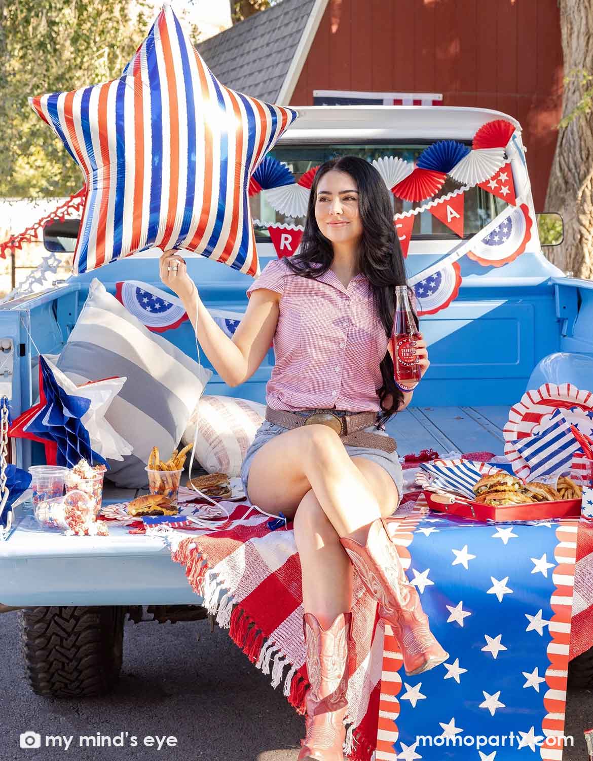A young lady in American-inspired outfits with red cowboy boots sitting on a light blue Ford truck bed with multiple USA, American Fourth of July-themed red, white and blue decorations from Momo Party including bunting garlands, red flag banners and American flag buntings, along with various USA themed tablewares, burgers and fries on a patriotic themed table runner. She is holding a large red, white and blue striped star shaped foil balloon in one hand while another hand is holding a coke bottle. 