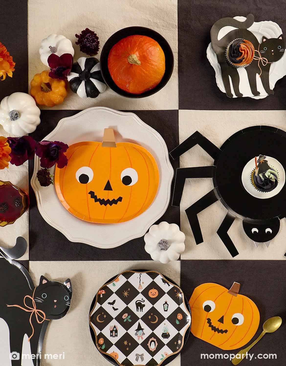 A styled Halloween table setting featuring Momo Party's Halloween themed party supplies including a pumpkin shaped plate with goggly eyes, a black cat shaped plate, a which shaped napkin and a black and white checkered pattern dessert plate on a black checkered tablecloth. Along with orange and white flowers, small pumpkin decorations in black, white and orange - making this a perfect look for a modern Halloween tablescape.
