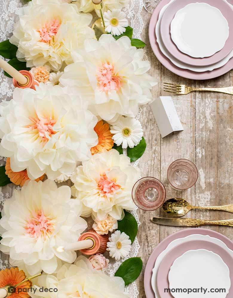 An elegant table set featuring layers of pink plates stacked and elegant gold utensils. For the centerpiece is Momo Party's cream flower pom pom decorations and blush candles. Making this a great inspiration for a rustic dinner table setting idea or wedding tablescape ideas.