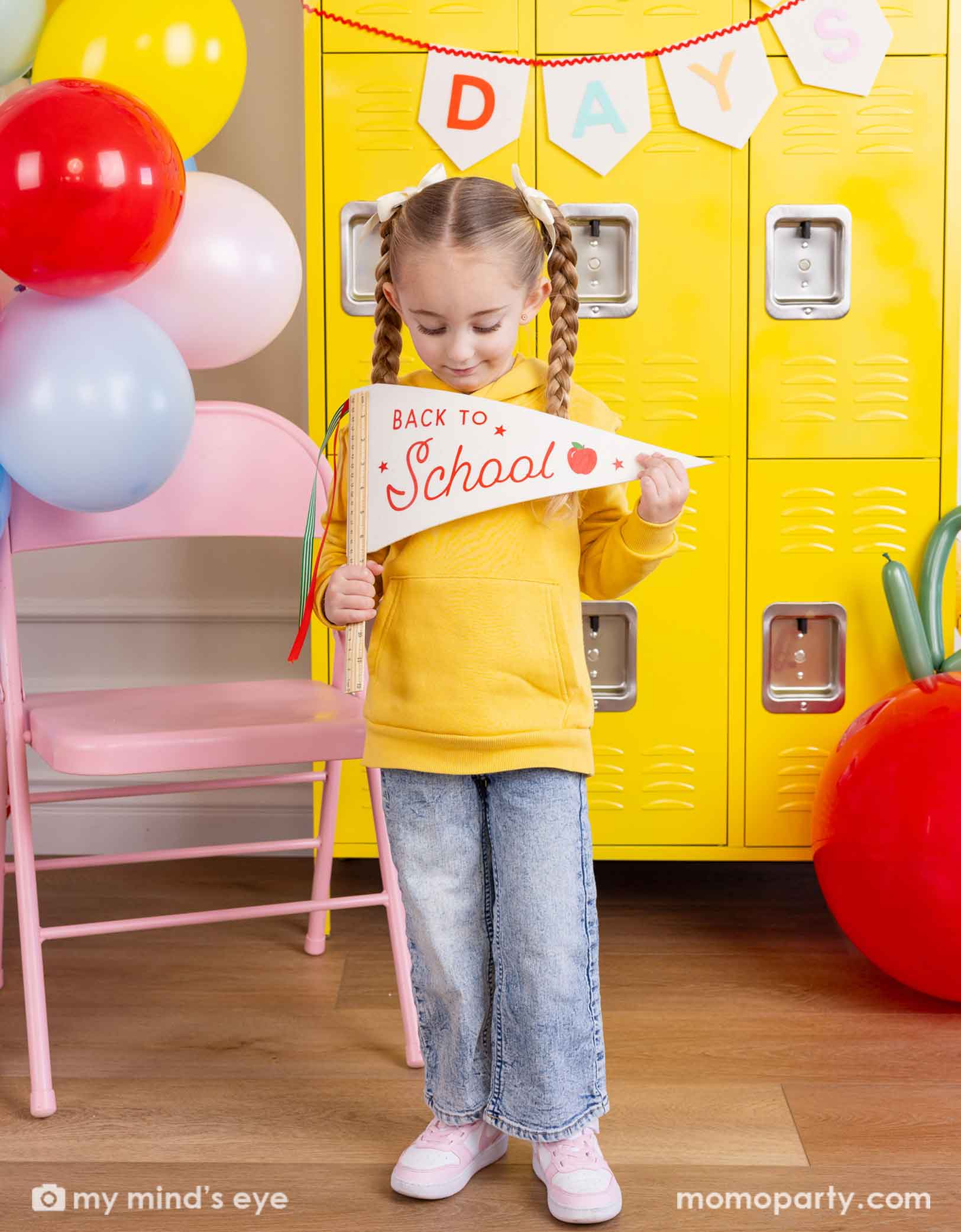 A girl in yellow shirt standing in front of a first day of school celebration decoration set up featuring various school themed party decors from Momo Party including School Days felt party banner and a festive balloon garland. She's holding the ruler back to school felt party pennant and looking at it. There's a yellow locker wall behind her and a pink folding chair. Making this a cute party decoration set up for kid's first day of school celebration.