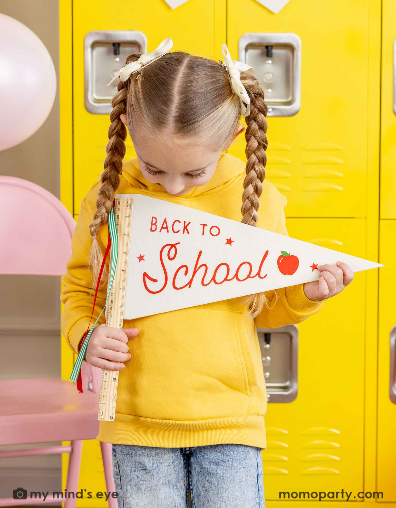 A girl in yellow shirt standing in front of a first day of school celebration decoration set up holding the ruler back to school felt party pennant and looking down at it. There's a yellow locker wall behind her and a pink folding chair. Making this a cute party decoration set up for kid's first day of school celebration.