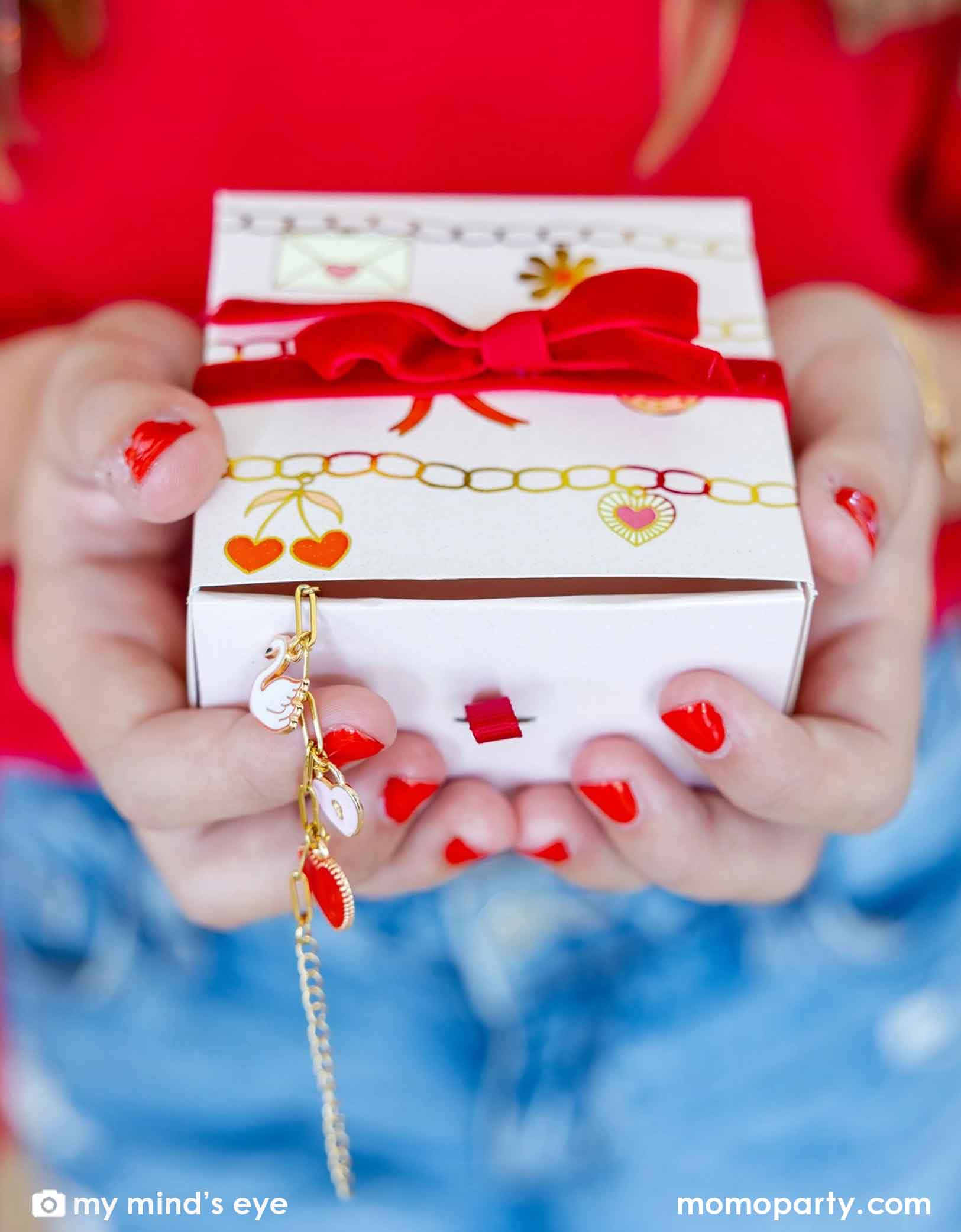 A girl with red nail polish is holding Momo Party's Valentine's Day Charm Bracelet Kit gift box with red ribbon and heart designs, accompanied by a gold bracelet.