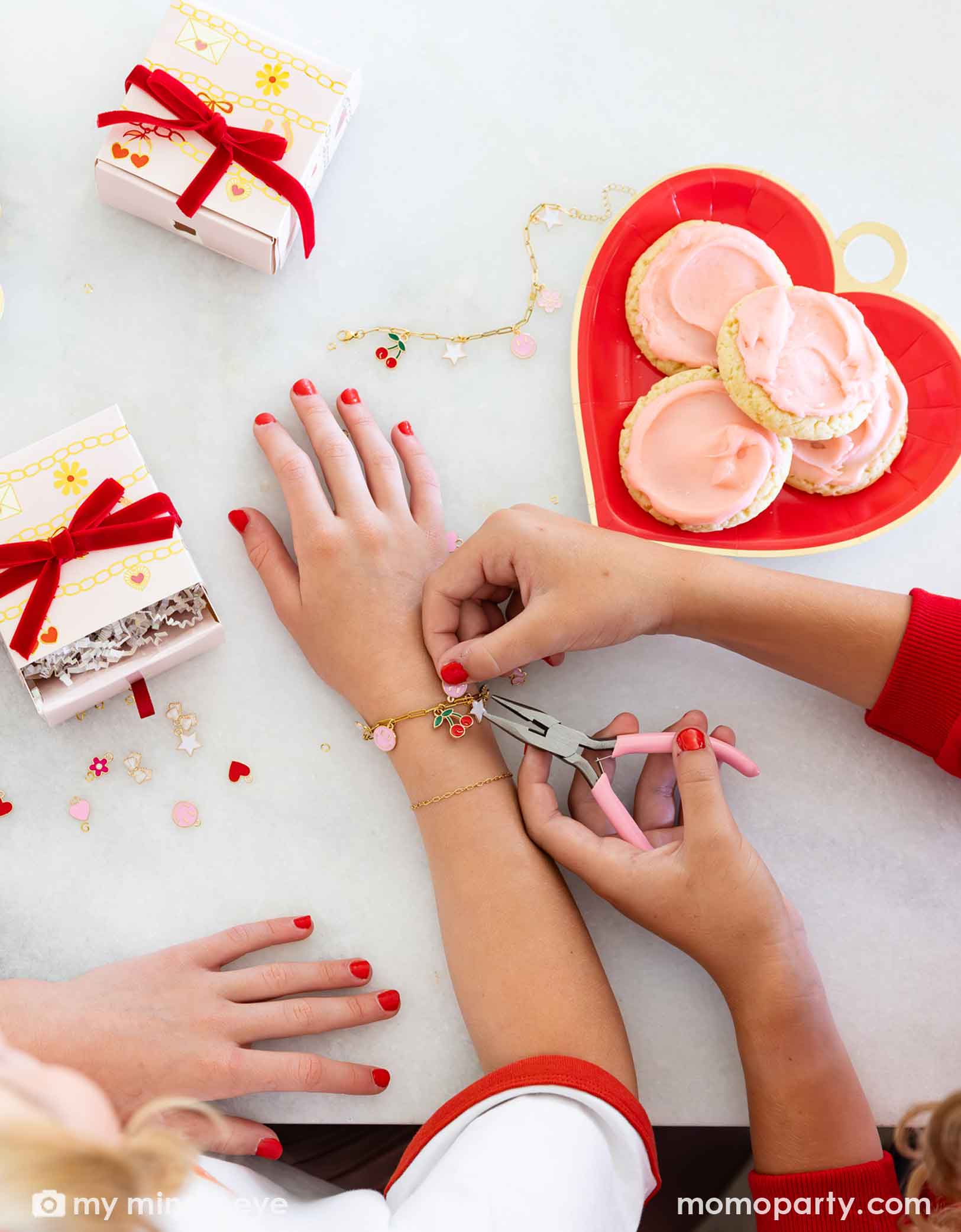 Children's hands with red nail polish getting their charm bracelets done with pink pliers, surrounded by small gift boxes and a heart-shaped plate of cookies at a Valentine's Day celebration.