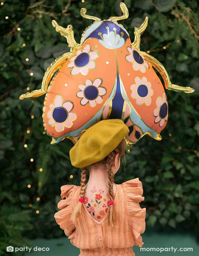 A little girl holding Momo Party's 34 x 30 inches ladybird shaped foil balloon by Party Deco. With coral color with small flower pattern with gold legs on it. She's dressed in her spring outfit with a mustard beret hat standing in a backyard with a wall filled of greenery.