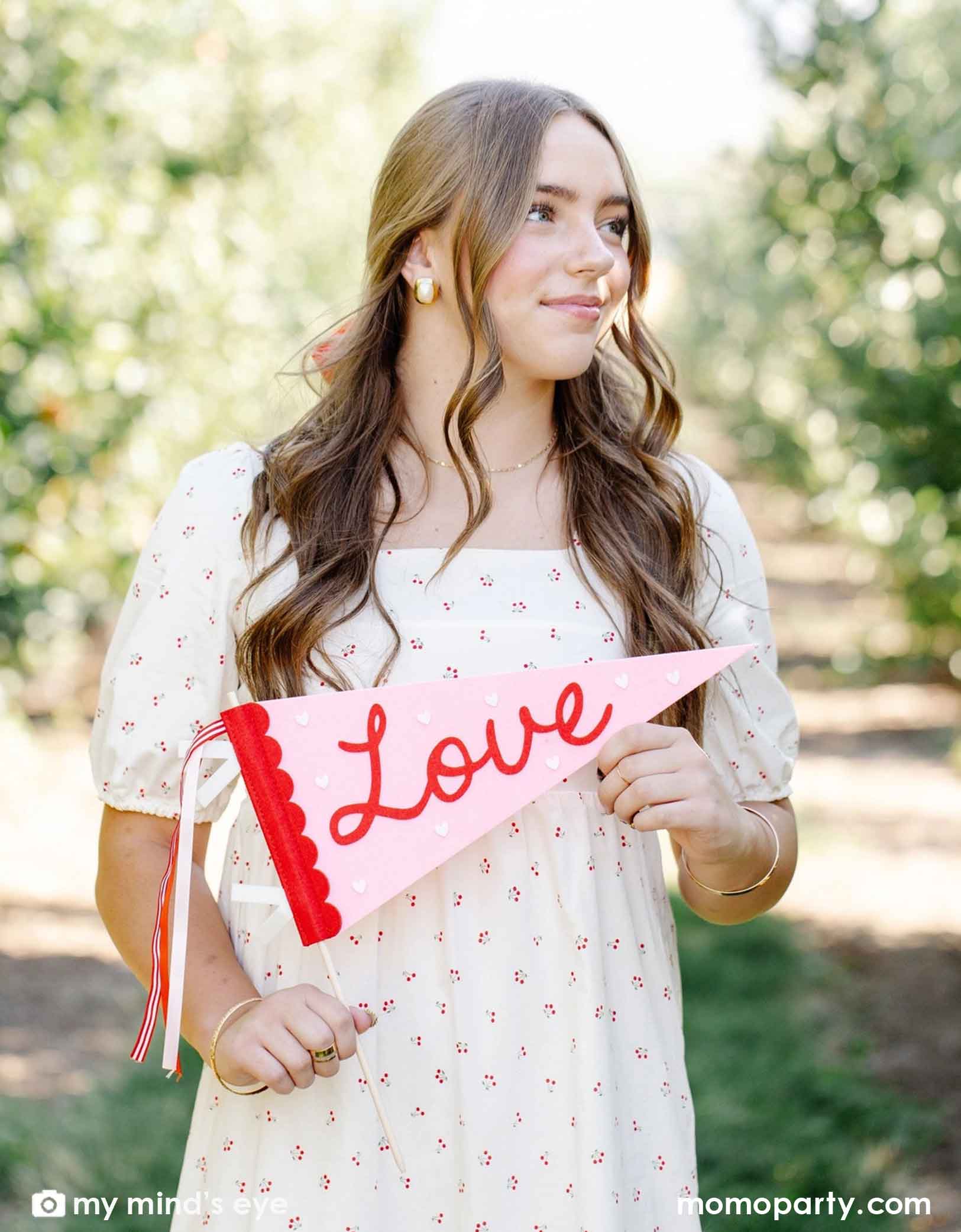 A girl in a white floral dress holding Momo Party's love felt party pennant standing in a open space with cherry trees behind.