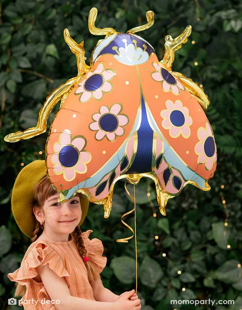 A little girl holding Momo Party's 34 x 30 inches ladybird shaped foil balloon by Party Deco. With coral color with small flower pattern with gold legs on it. She's dressed in her spring outfit with a mustard beret hat standing in a backyard with a wall filled of greenery.