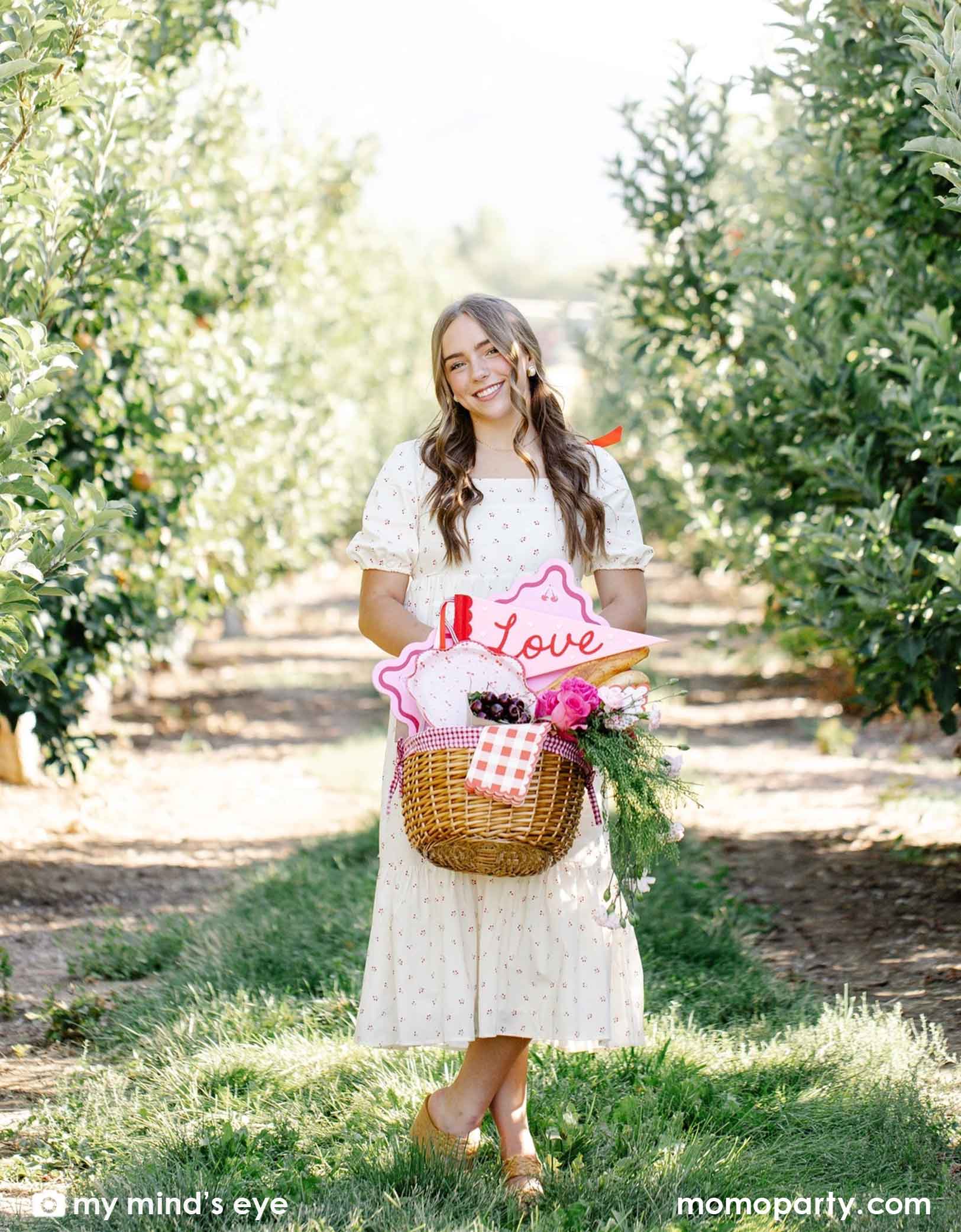 A girl in a white floral dress carrying a basket full of flowers and cherry themed party supplies from Momo Party including plates, placemats, napkins, and the love felt party pennant. She is standing in a open space with trees behind her.