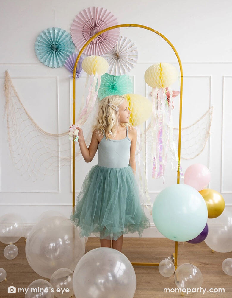 A young girl in her teal tutu dress standing in front of a white wall decorated with mermaid inspired party decorations including Momo Party's under the sea paper fans in pastel colors of aqua, pink, lilac, teal, cream and peach. Next to the paper fans hung a fishing net draping over a golden arch structure adorned with honeycomb jelly fish decorations by My Mind's Eye. On the floor there are some clear latex balloons as bubbles, making this an inspo for an enchanting kid's mermaid birthday party set up.