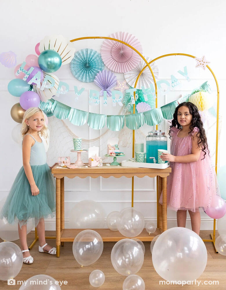 Two school-aged girls in an enchanting kid's mermaid birthday party set up featuring mermaid inspired decorations and balloons from Momo Party including the Under the Sea paper fan set in pastel colors, Mermaid Happy Birthday Banner Set and shell shaped paper decorations by My Mind's Eye on the wall. The girls are standing by a party table with mermaid inspired party supplies including cups, plates, napkins and treat boxes. On the floor are some clear latex balloons creating a whimsy water bubble scene.