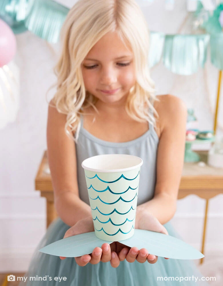 A school-aged girl in a teal tutu dress holding Momo Party's mermaid tail paper cups by My Mind's Eye. In her back you can see some mermaid party decorations on the wall behind a party table featuring teal and aqua themed party supplies for a under the sea/mermaid birthday celebration.