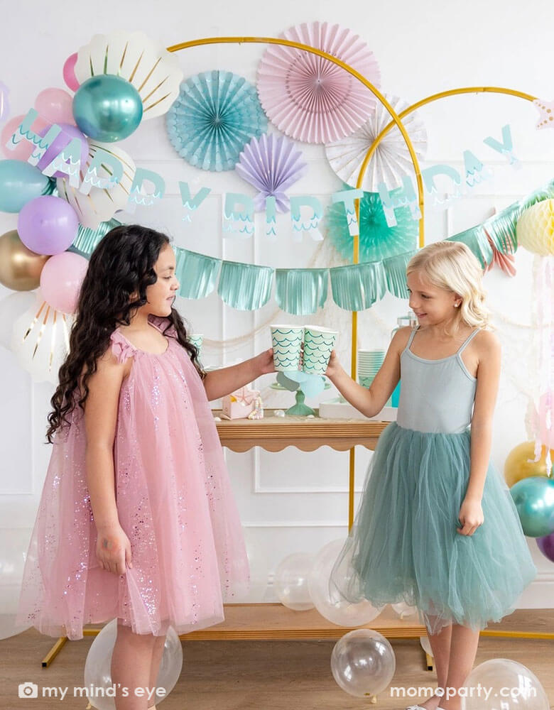 Two school-aged girls in their teal and pink tutu dresses celebrating a birthday in an enchanting kid's mermaid birthday party set up featuring mermaid decorations and balloons from Momo Party including the Under the Sea paper fan set in pastel colors of light pink, aqua, teal, lilac and cream, Mermaid Happy Birthday Banner Set with aqua foil fringe banner and shell shaped paper decorations by My Mind's Eye on the wall. The girls are raising their aqua mermaid tail party cups and cheering to each other.