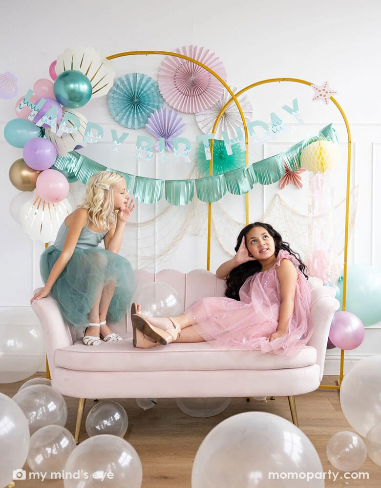 Two school-aged girls sitting on a shell shaped blush sofa in an enchanting kid's mermaid birthday party set up featuring mermaid inspired decorations and balloons from Momo Party including the Under the Sea paper fan set in pastel colors of light pink, aqua, teal, lilac and cream, Mermaid Happy Birthday Banner Set with aqua foil fringe banner and shell shaped paper decorations by My Mind's Eye on the wall. On the floor are some clear latex balloons in various sizes creating a whimsy water bubble scene.