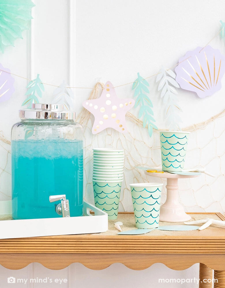 A mermaid party table features Momo Party's mermaid tail paper cups by My Mind's Eye. Next to the paper cups is a water dispenser filled with aqua colored drink. The table is decorated with shells, sea star and white sand decorations. In the back there's a fishing net hung on the wall which is adorned with under the sea paper party pennants features sea stars, sea grass, and sea shells in soft pastel colors, making this a perfect inspo for a kid's mermaid, under the sea party decorations.
